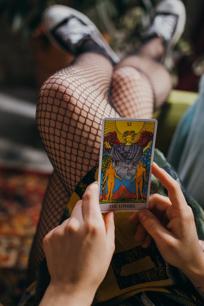 Close-up of hands holding 'The Lovers' tarot card, with fishnet stockings visible.